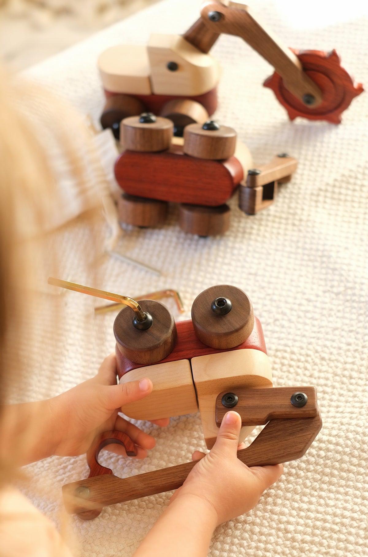 Child's hands taking apart the Maple & Padauk Excavator with an Allen key on a white throw, with the trench digger and another wooden vehicle behind