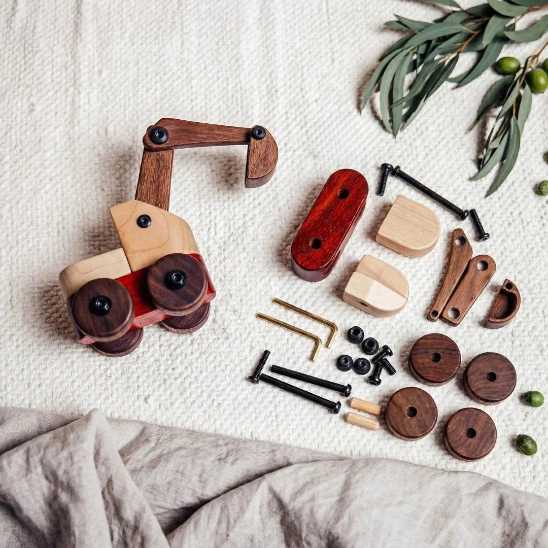 Maple & Padauk Excavator assembled beside its loose maple, padauk and walnut parts, bolts and two Allen keys, on a cream throw with olive sprigs and a linen cloth
