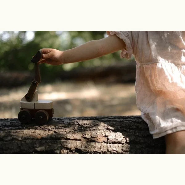 Child in linen dress playing with Maple & Padauk Excavator on a fallen log outdoors, maple body and walnut boom visible