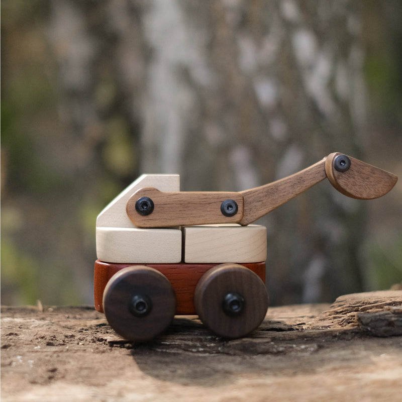 Maple Padauk Walnut excavator toy on weathered log outdoors, showing pale cream, coral-red, and dark brown hardwood tones