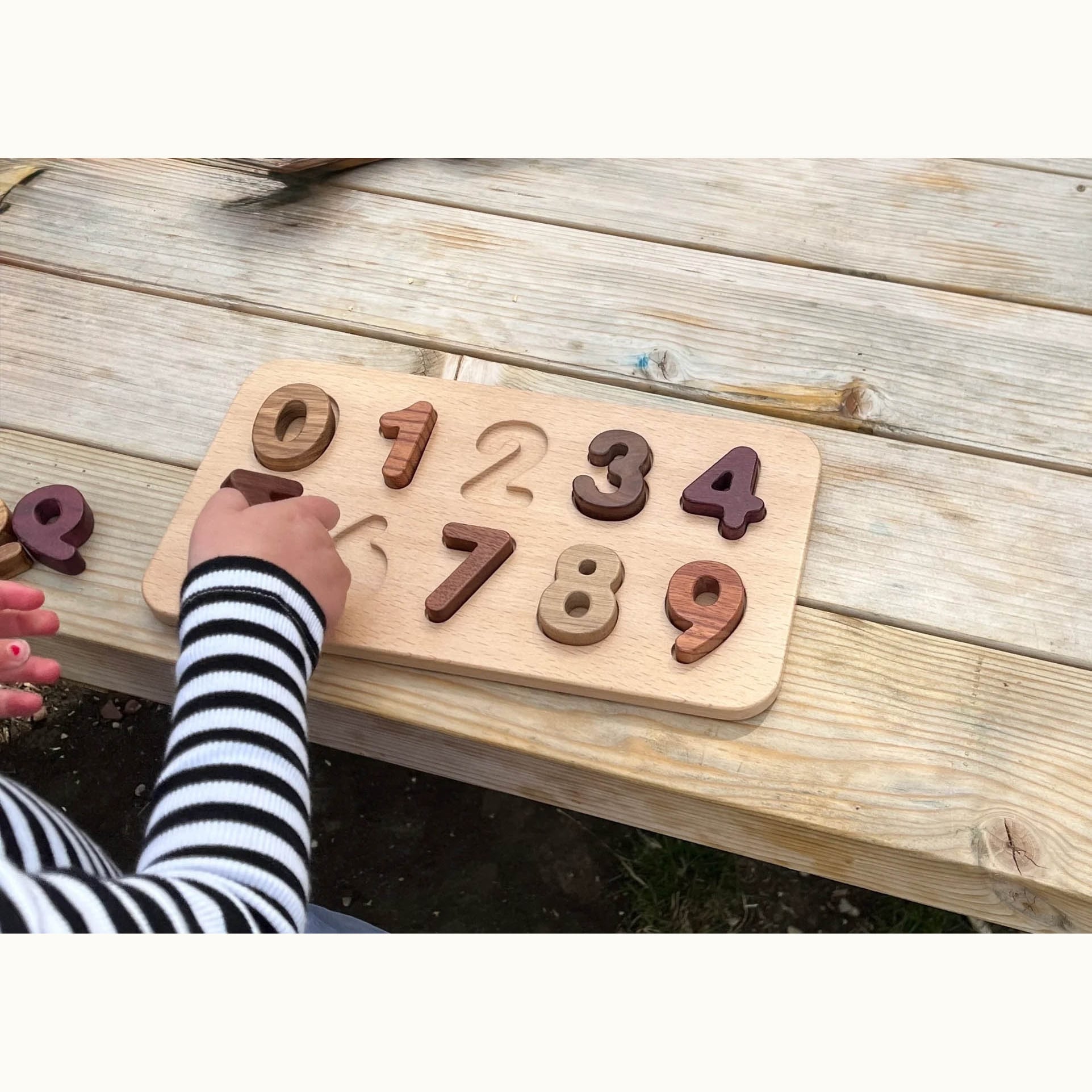 Child placing Walnut, Purpleheart & Tigerwood number puzzle pieces on beech board outdoors, digits 0–9 in natural hardwood to