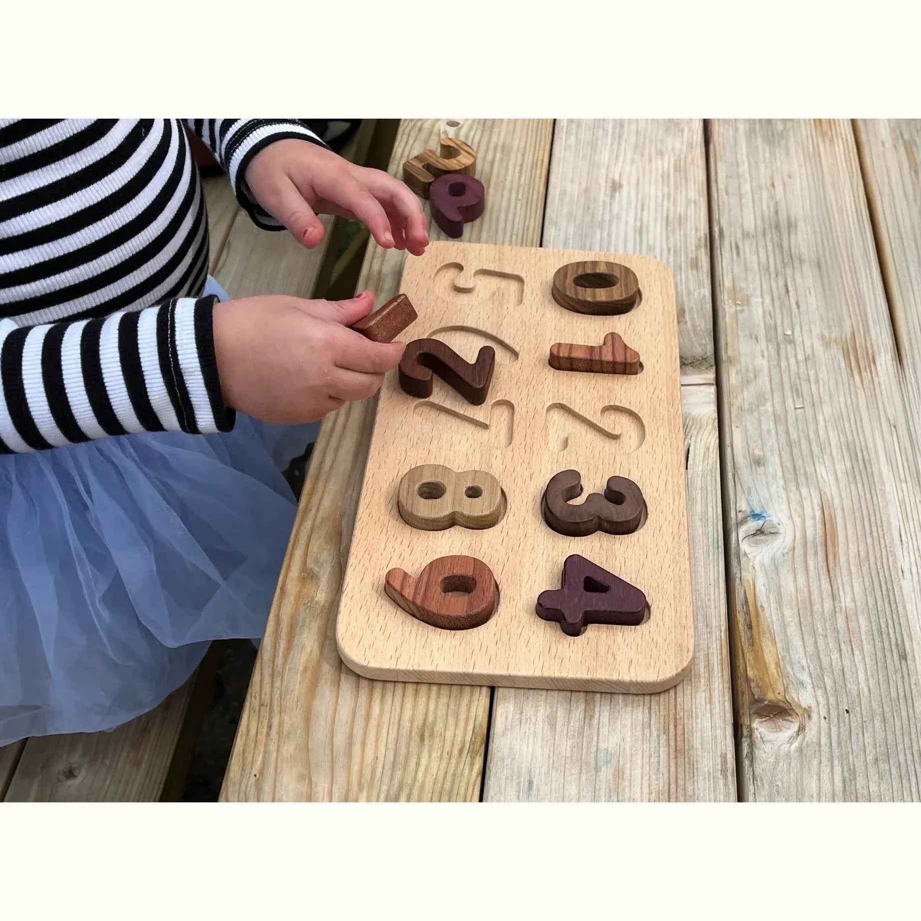 Toddler assembling Oak & Sprout wooden number puzzle outdoors, showing walnut, purpleheart and tigerwood pieces in slots 0–8