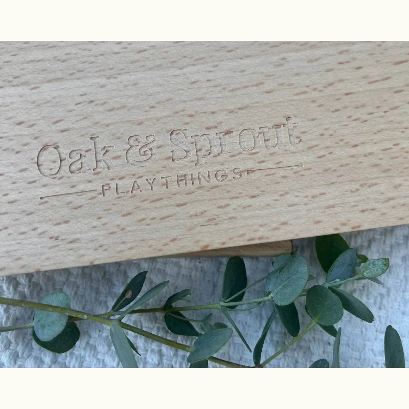 Back of Oak & Sprout Wooden Number Puzzle showing laser-engraved brand logo on pale hardwood, with eucalyptus sprig on linen