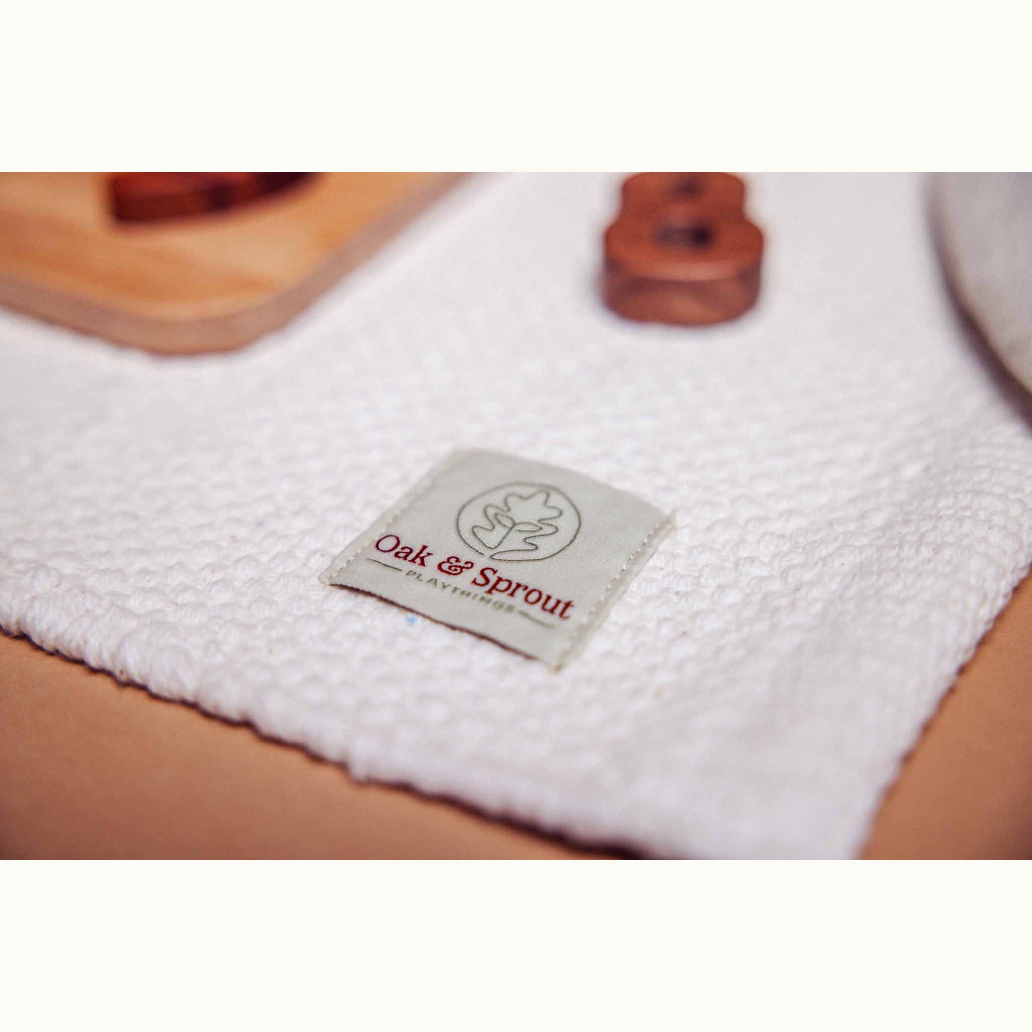 Oak & Sprout Playmat woven brand label on white fabric, with walnut and maple toy pieces softly visible behind