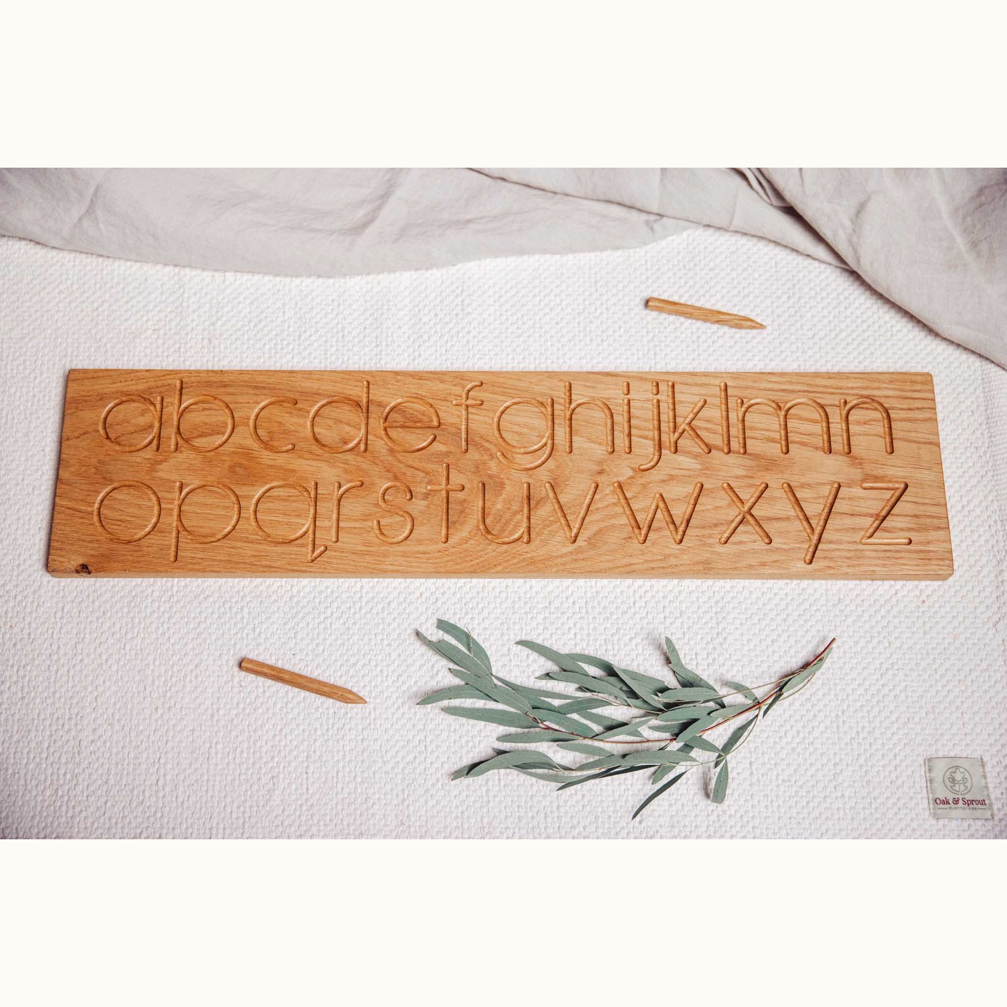 Oak alphabet tracing board flat-lay showing all 26 routed lowercase letters in two rows, with wooden stylus on linen