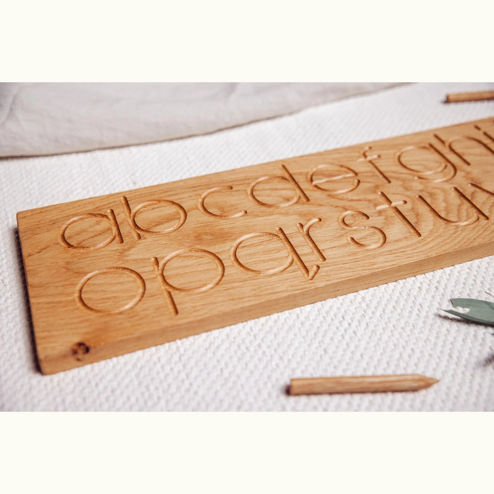 Oak alphabet tracing board close-up showing carved lowercase letter grooves a–u with oak stylus tools on white muslin