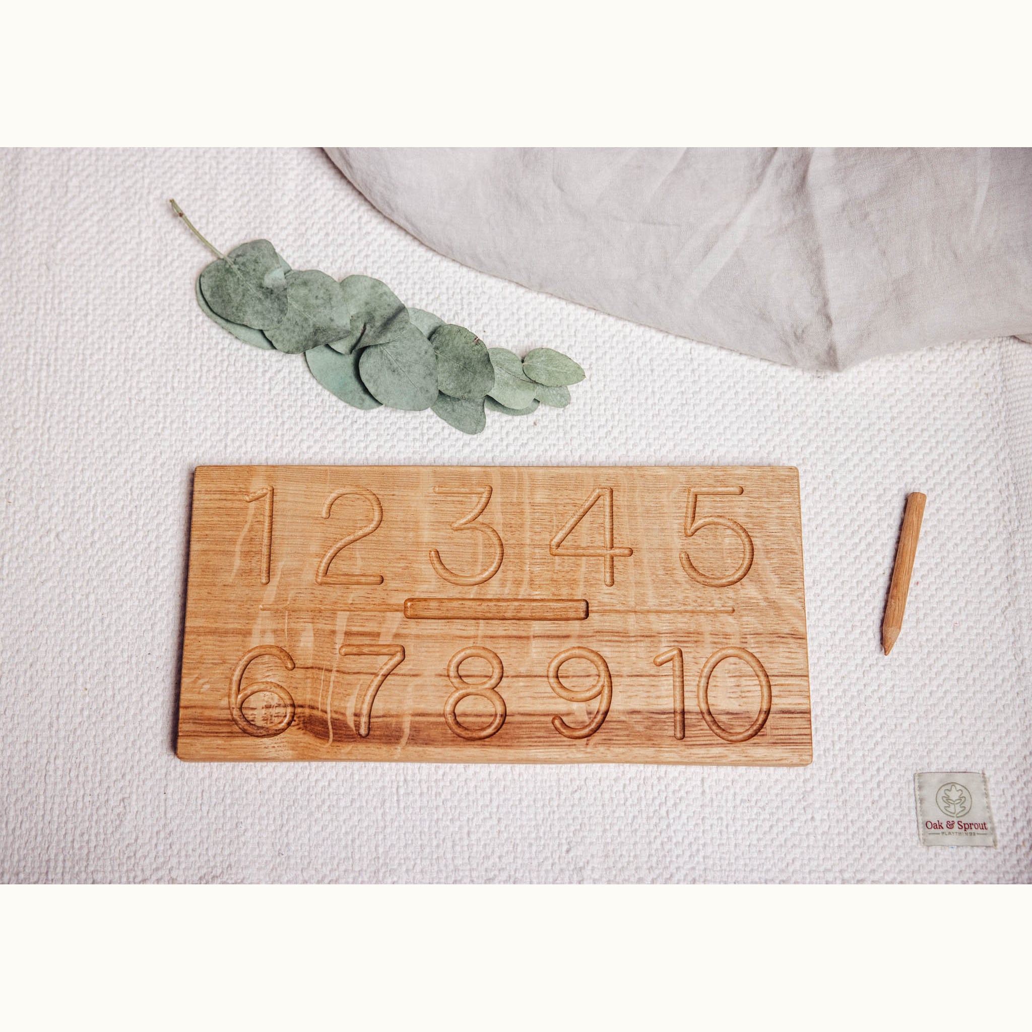 Oak Numbers Tracing & Balls board flatlay showing carved 1–10 numeral grooves, styled with eucalyptus sprig and wooden stylus