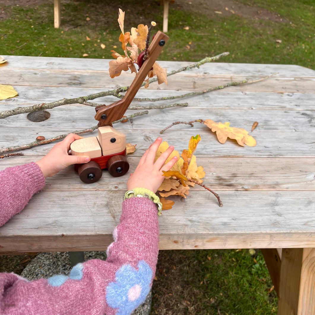 Child playing with wooden toy truck and leaves on a wooden table outdoors.