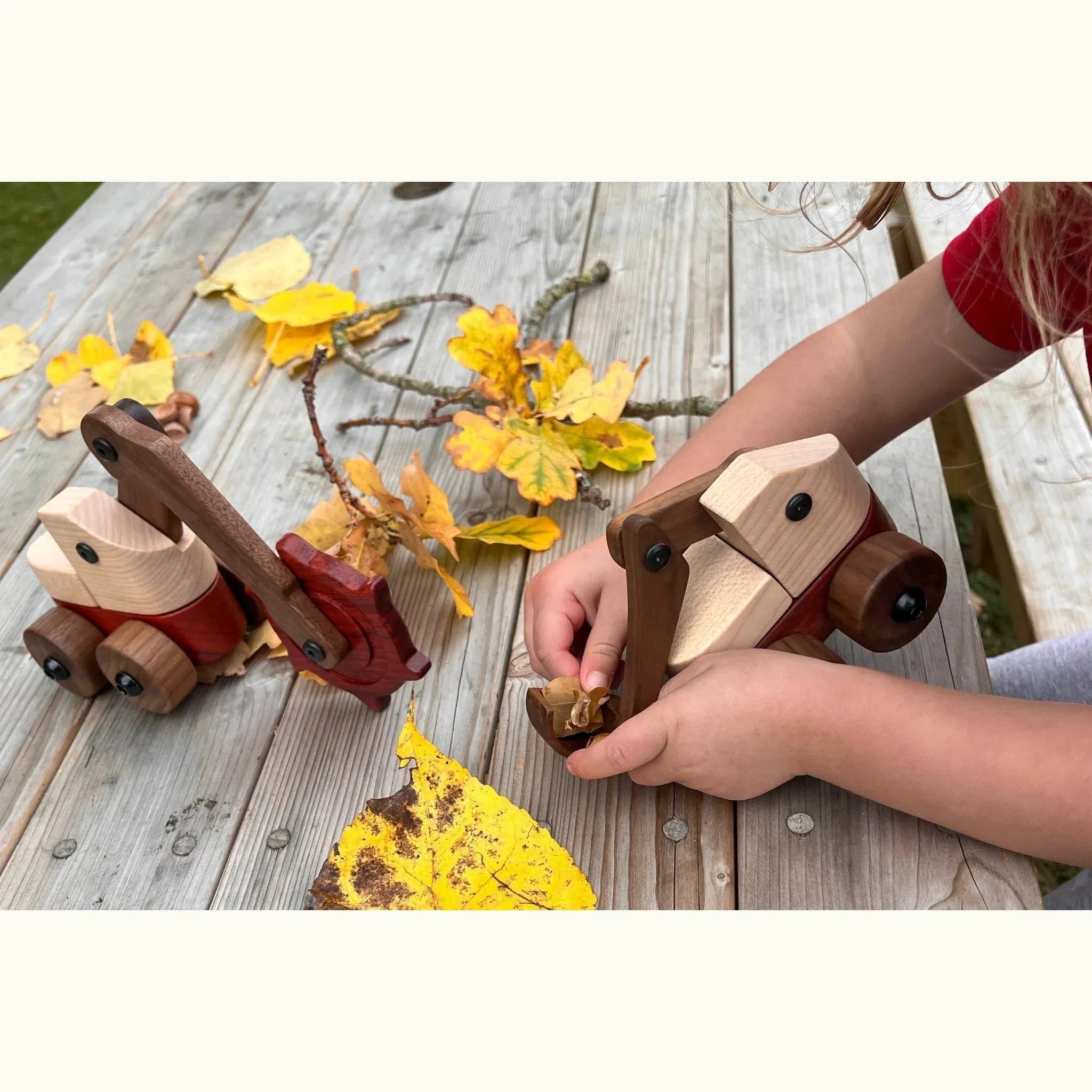 Child playing with two maple, padauk and walnut excavator toys on an outdoor deck surrounded by autumn leaves