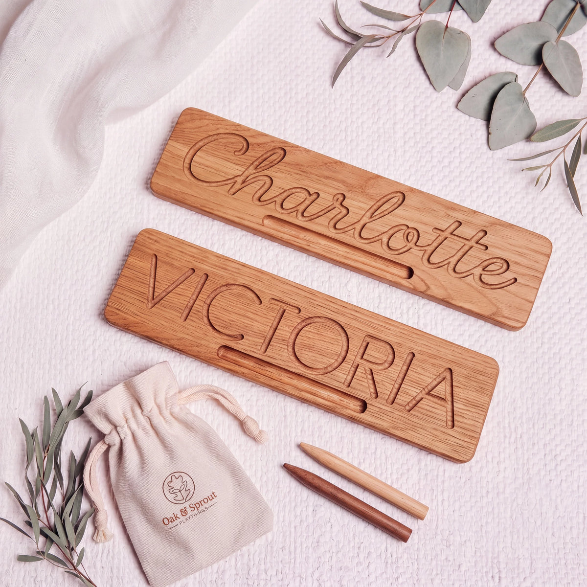 Two oak Name Tracing Boards flat-lay: cursive 'Charlotte' and block 'VICTORIA', with stylus tools and cotton pouch