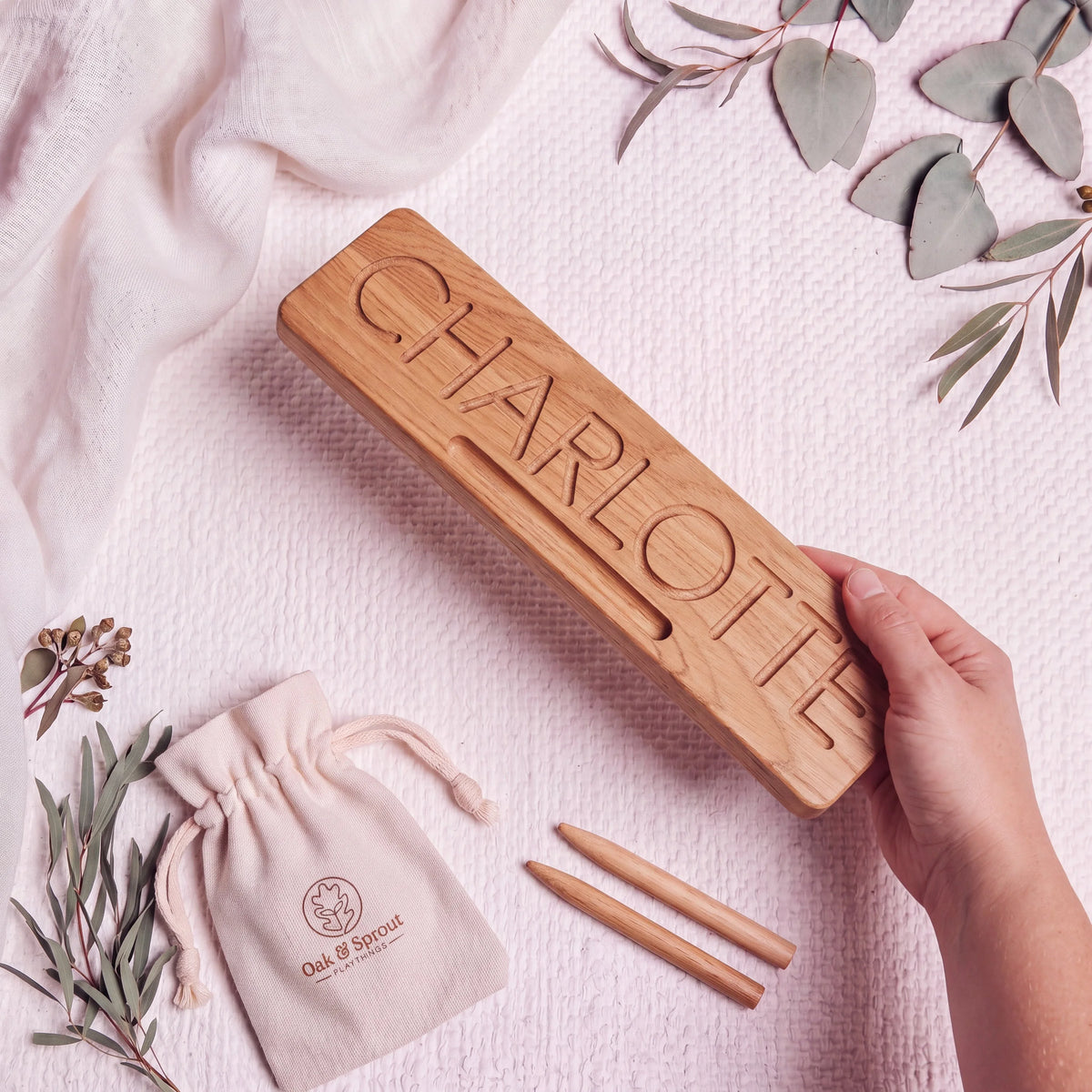 Oak name tracing board engraved with CHARLOTTE, held at angle with two styluses and branded cotton gift pouch on blush
