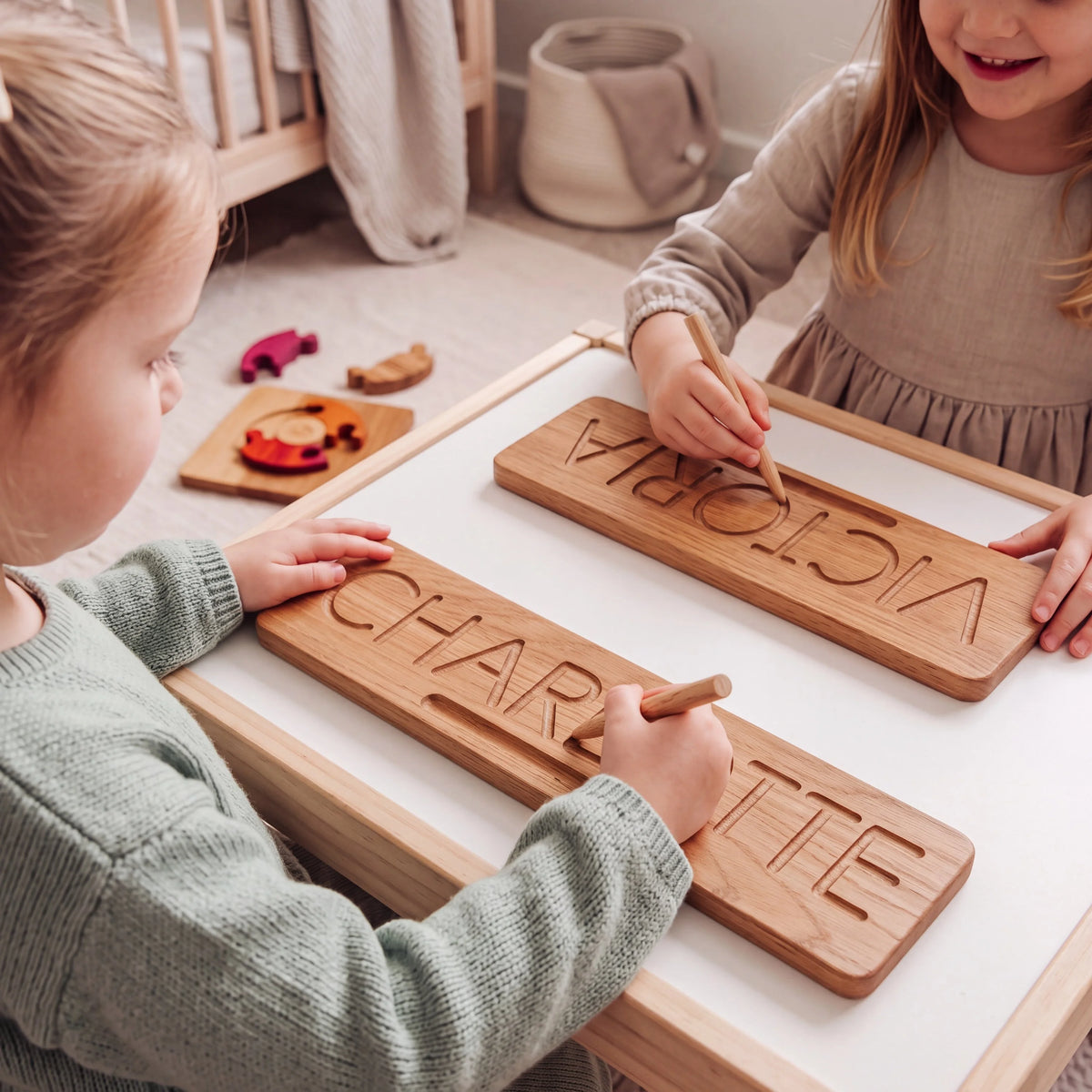 Two children tracing grooved oak name boards reading CHARLOTTE and VICTORIA with wooden styluses at a nursery table