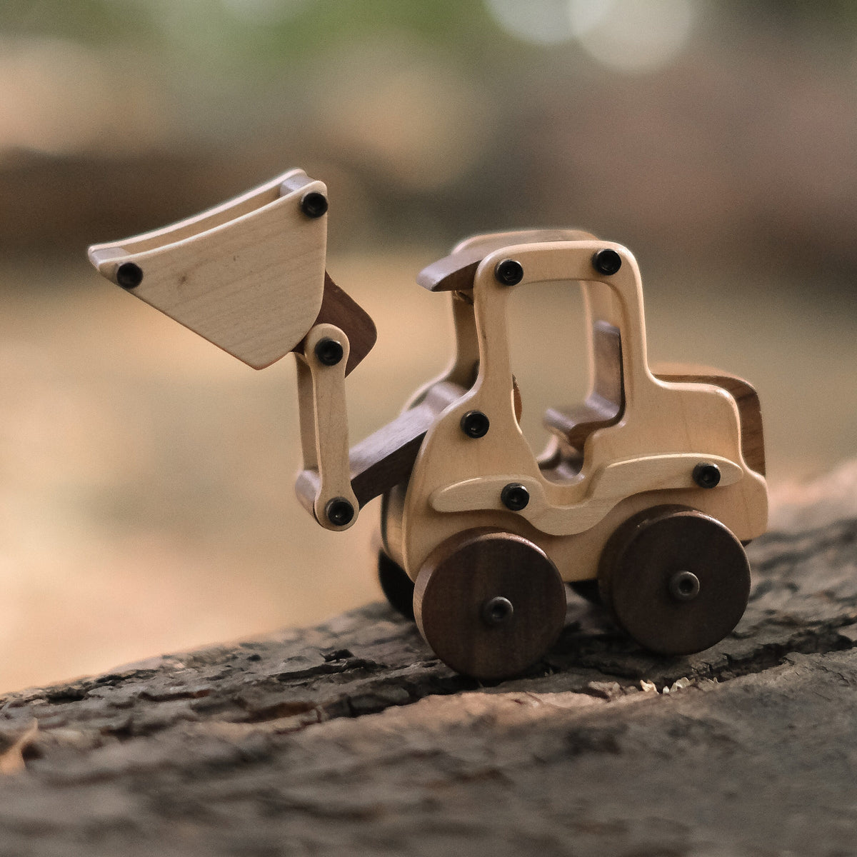 Maple Digger toy on tree bark outdoors, pale maple cab and arm, dark walnut wheels, bucket raised, metal bolts visible