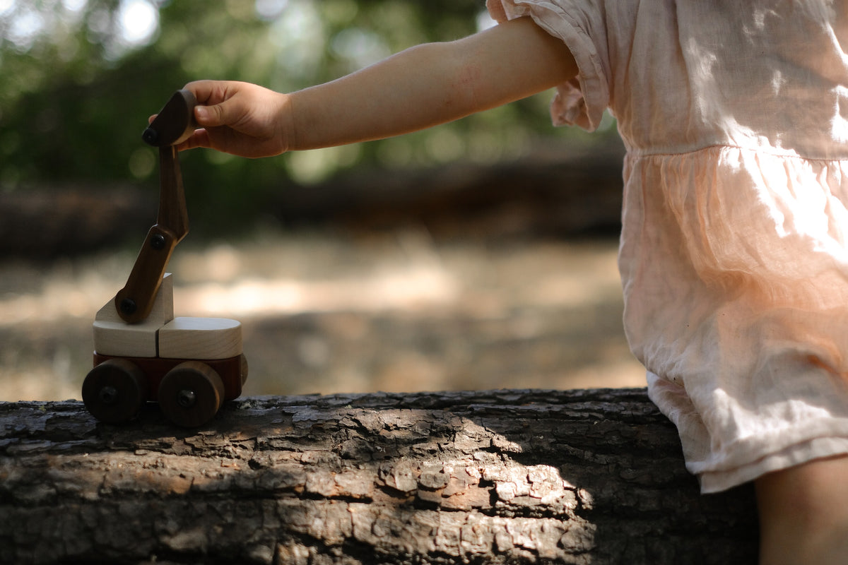 Child in a pink linen dress moving the arm of the Maple & Padauk Excavator on a fallen log in the woods