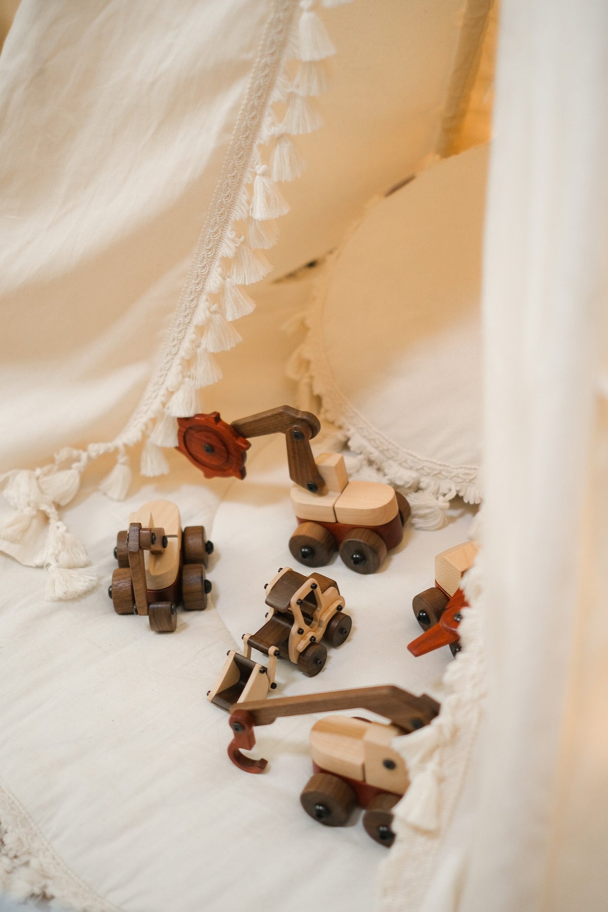 Walnut & Maple Crane in the foreground with other Oak & Sprout wooden vehicles, including the trench digger and Maple Digger, on cream fabric in a tasselled play tent