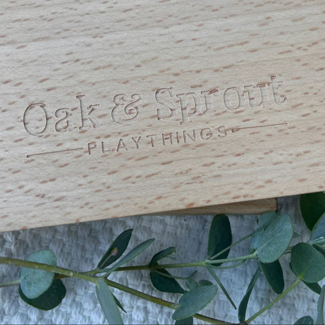 Wooden box with 'Oak & Sprout Playthings' engraving, surrounded by green leaves.
