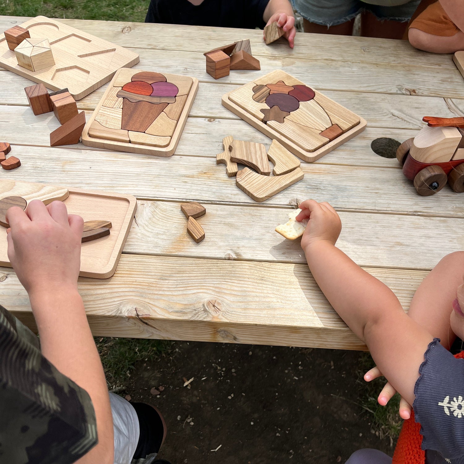 Children playing with wooden puzzle toys on a wooden table outdoors.