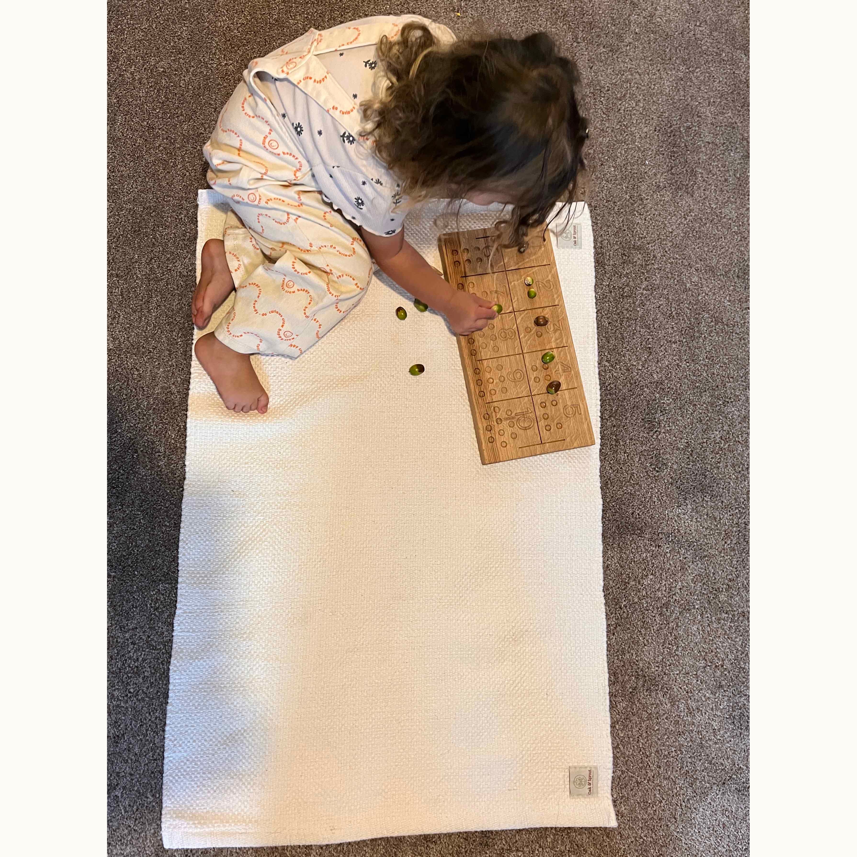 Child sitting cross-legged on Oak & Sprout Playmat using a walnut counting board with green beads, bird's-eye view