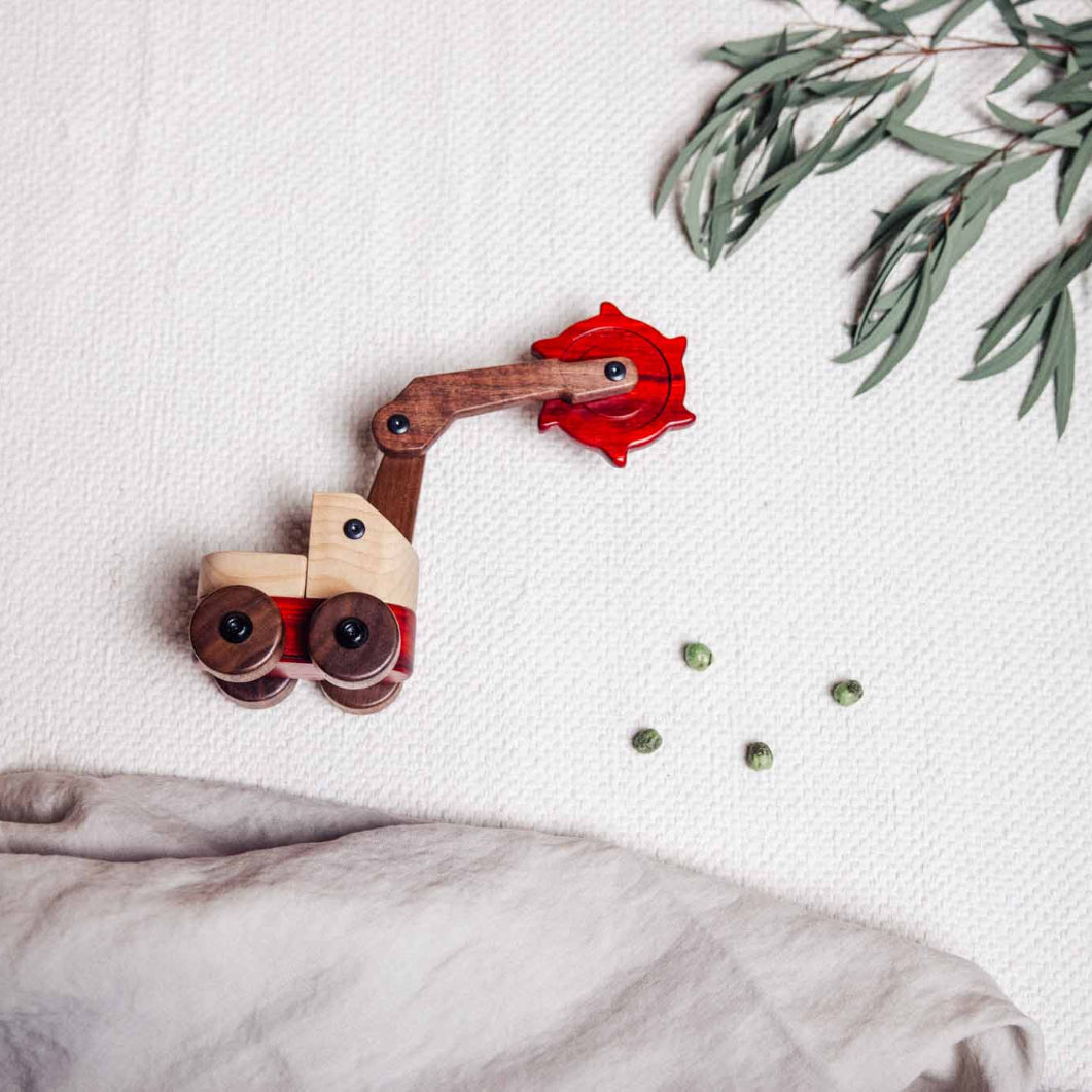 Wooden toy bulldozer with red wood on a white surface with green leaves