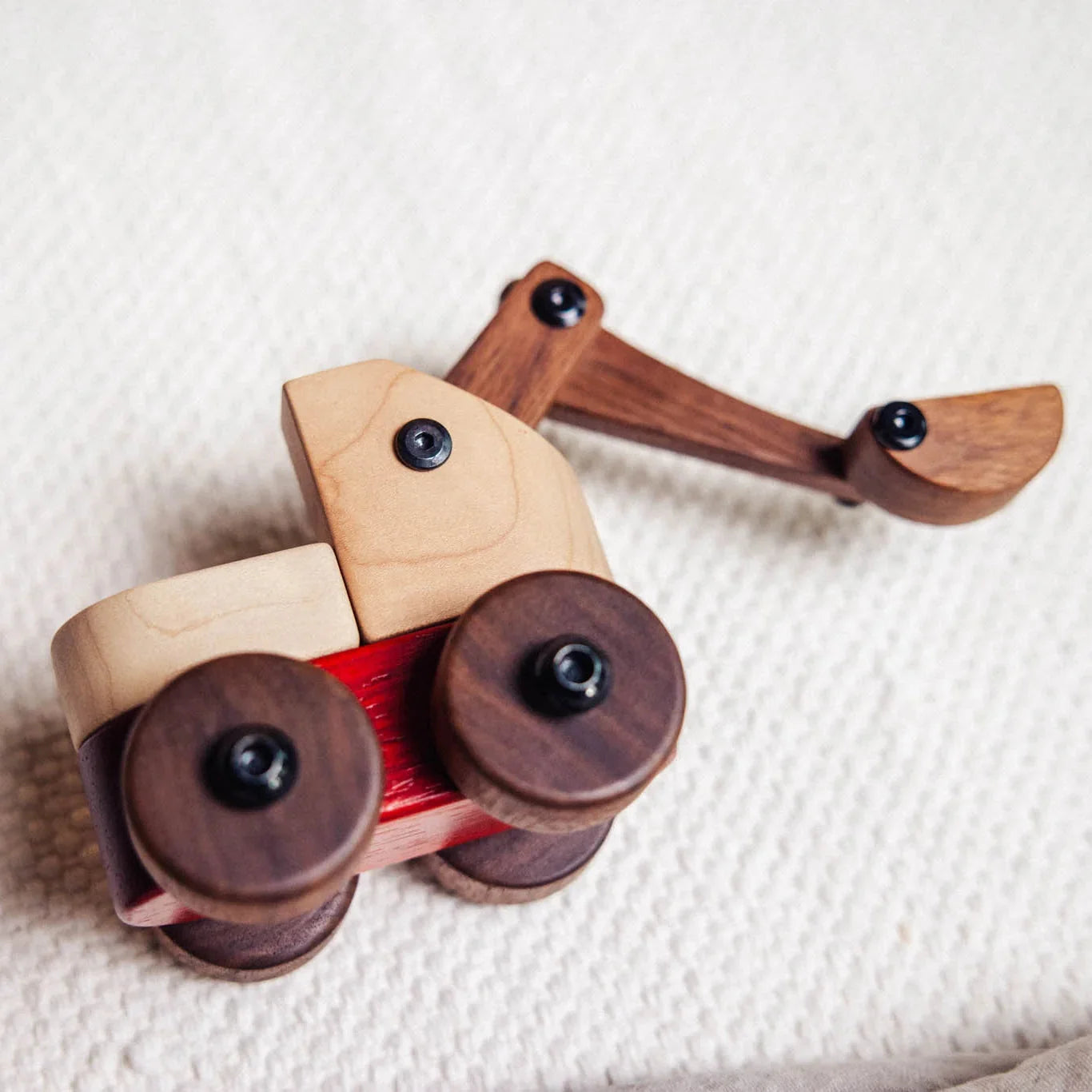 Maple Padauk Walnut excavator toy close-up on knit fabric, showing coral Padauk chassis, cream Maple cab, dark Walnut wheels 
