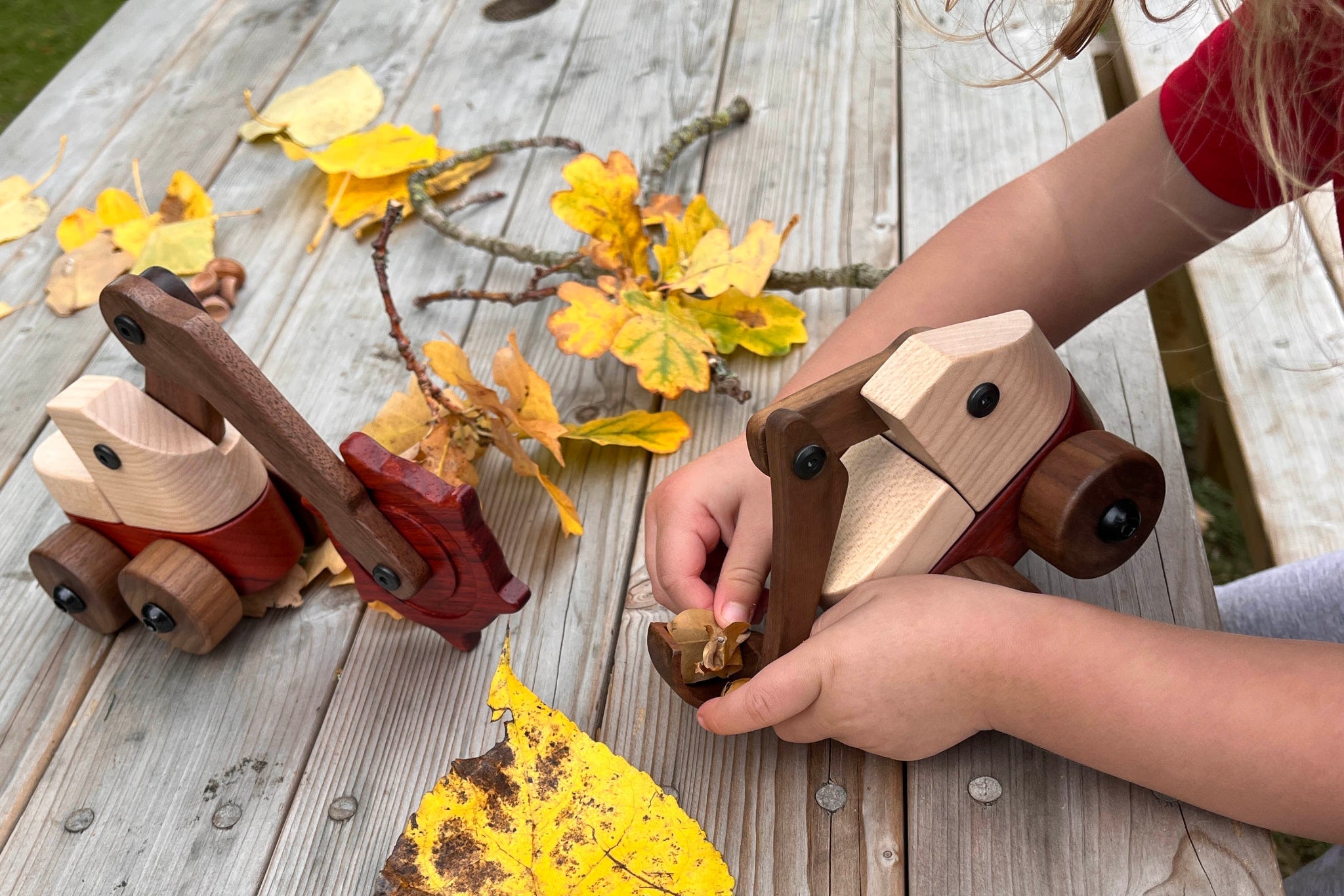 Child playing with wooden toy diggers on a wooden surface with autumn leaves.