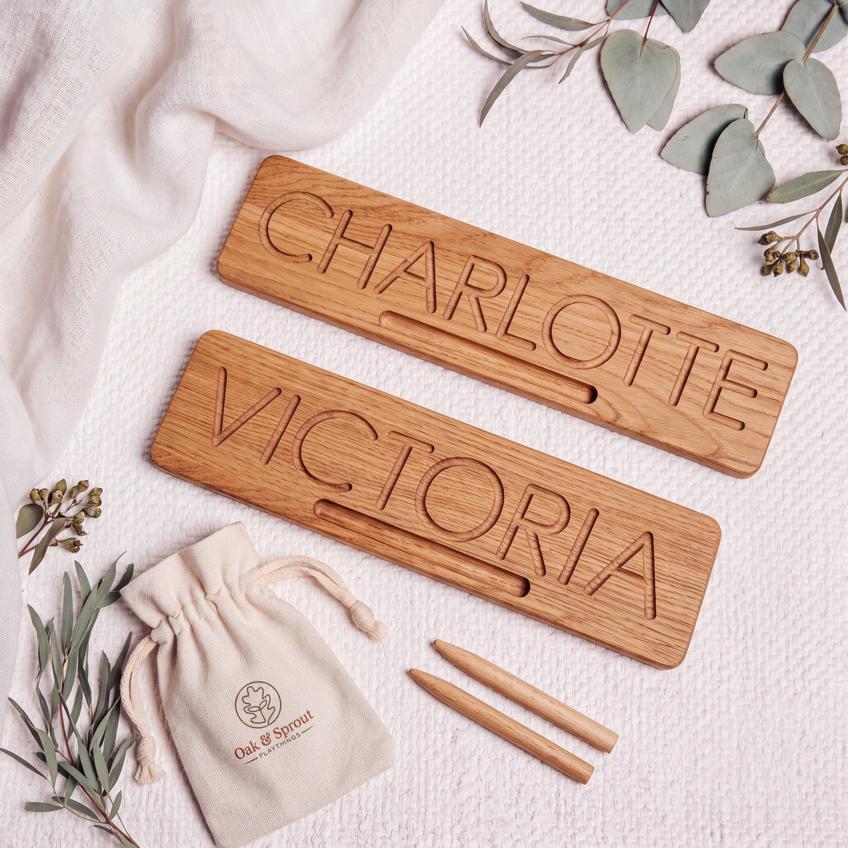 Two personalised oak Name Tracing Boards engraved with CHARLOTTE and VICTORIA, shown with stylus tools and cotton pouch on