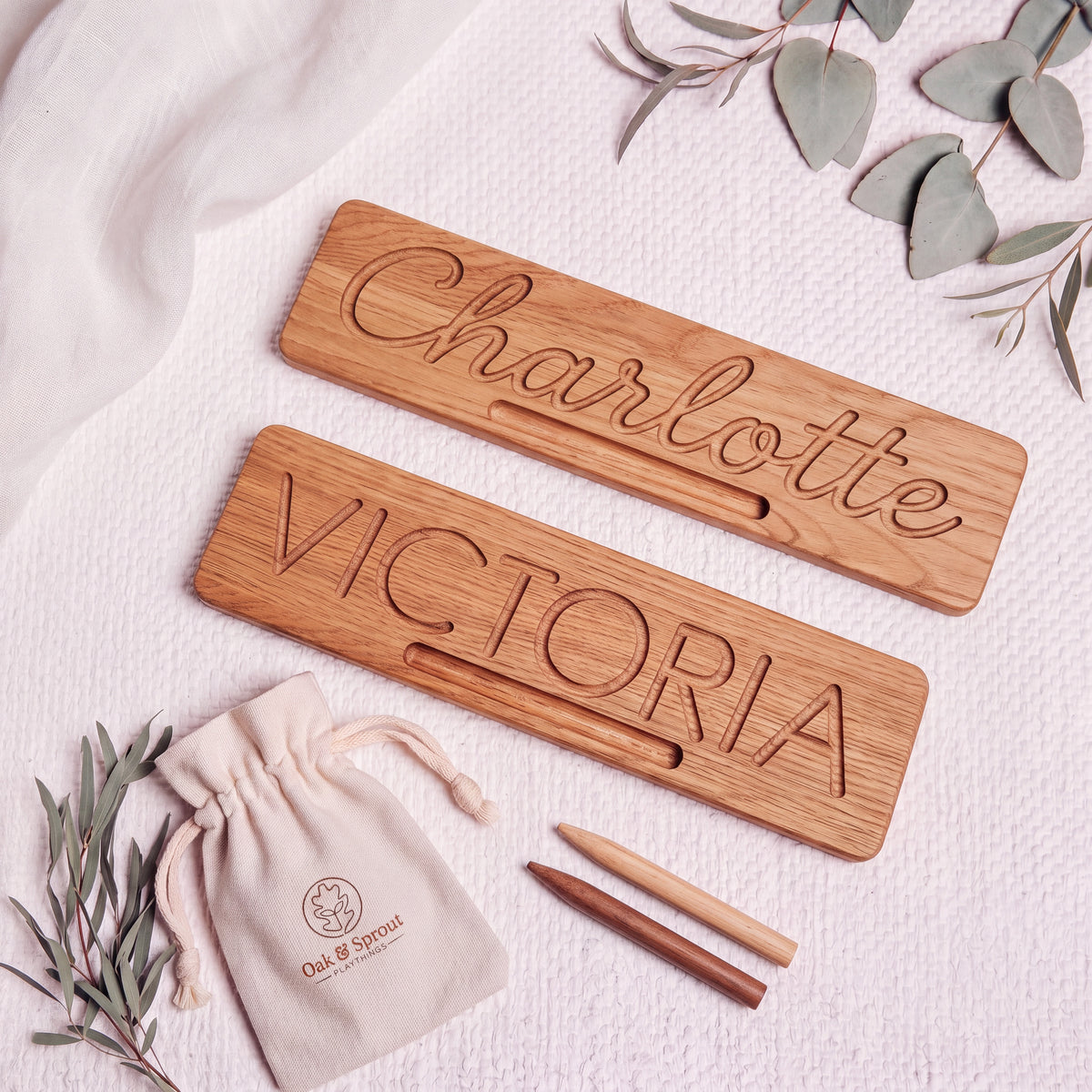 Two oak Name Tracing Boards flat-lay: cursive 'Charlotte' and block 'VICTORIA', with stylus tools and cotton pouch