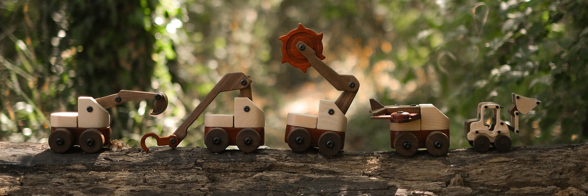 Five handcrafted hardwood construction vehicles — crane, excavator, mixer, loader — displayed on a log in maple, walnut, and
